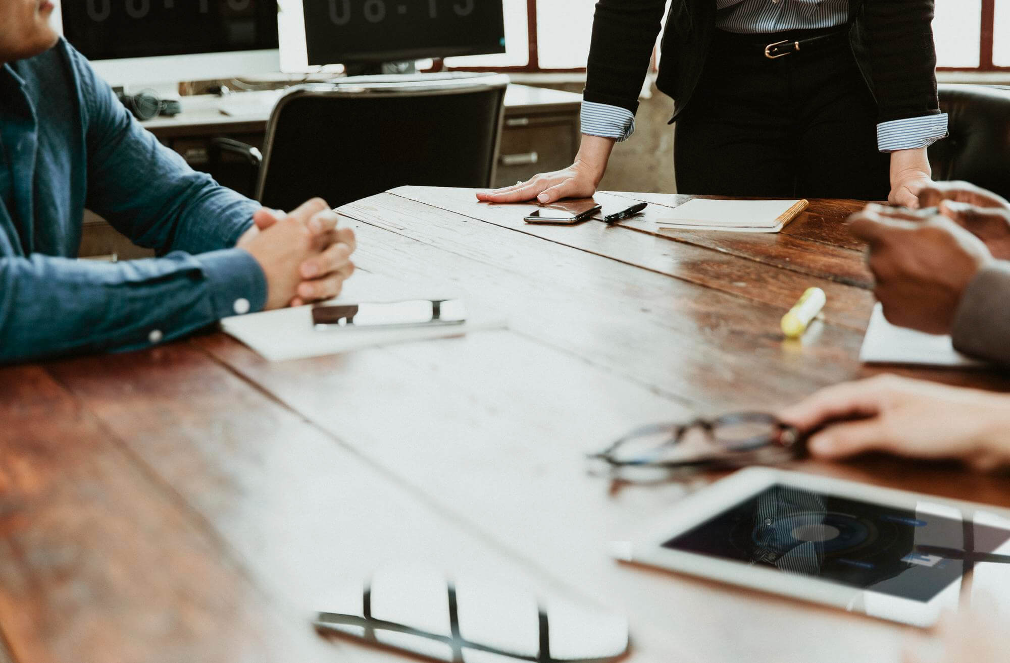 Kokoushuoneen puupöydällä on ihmisiä, muistivihko, älypuhelin, silmälasit ja tabletti, valaistus on kirkas.